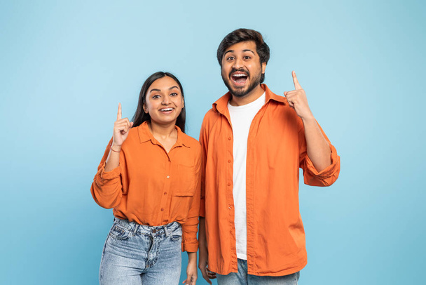 Ινδός άνδρας και γυναίκα και οι δύο σε πορτοκαλί, ενθουσιασμένοι δείχνοντας επάνω με τις φωτεινές εκφράσεις σε ένα μπλε υπόβαθρο - Φωτογραφία, εικόνα