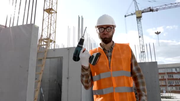 Opravář v uniformě a helmě drží bezdrátový šroubovák, stojí na svém pracovišti - Záběry, video
