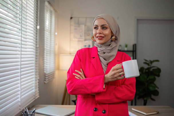 Εμπιστευτική μουσουλμάνα επιχειρηματίας στέκεται μπροστά από το γραφείο στο γραφείο. - Φωτογραφία, εικόνα