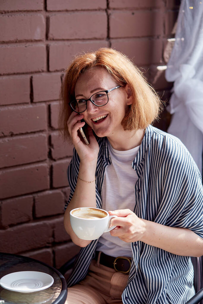 Счастливая улыбающаяся женщина в очках разговаривает по телефону во время кофе-брейка. - Фото, изображение