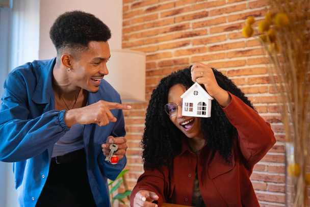 Mężczyzna z kluczami wskazującymi na mini dom trzymający jego dziewczynę z jej nowego domu, pozujący szczęśliwie przed kamerą.. - Zdjęcie, obraz
