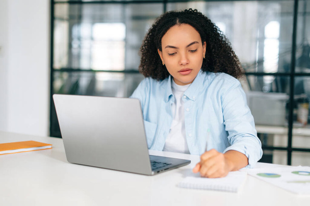 Soustředěný chytrý zaneprázdněný brazilský nebo hispánský kudrnatý žena v pastelové modré košili, manažer, sekretářka, sedí v moderní kanceláři u pracovního stolu, pracuje na notebooku, dělá poznámky do notebooku, plánování - Fotografie, Obrázek