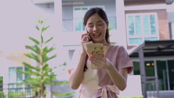 Portret szczęśliwej Azjatki trzymającej przed domem dużo banknotów, by uczcić i spłacić ostatnią ratę domu, Pożyczki na nieruchomości, Uśmiechnięty młody sukces - Materiał filmowy, wideo