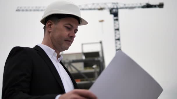 Architect studying details on paper drawings in his hands in the background of a construction crane and an unfinished building. Architect in business attire and white helmet action on the street - Footage, Video