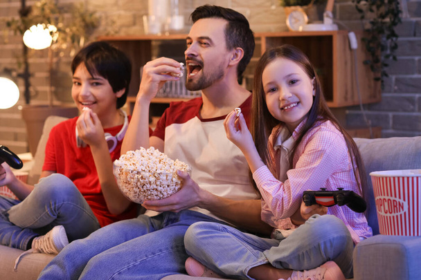 Ojciec i jego małe dzieci jedzą popcorn podczas gry wideo w domu wieczorem - Zdjęcie, obraz