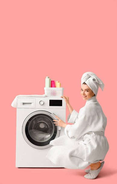 Jovem mulher em roupão de banho com máquina de lavar roupa sobre fundo rosa - Foto, Imagem