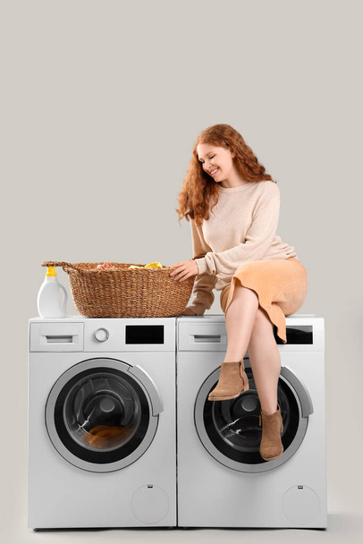 Young woman with laundry basket and washing machines on light background - Фото, зображення