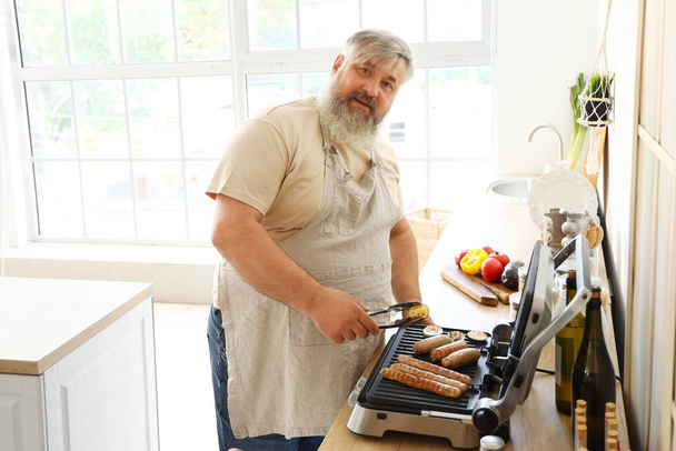 Hombre maduro con tong cocinar sabrosas salchichas y verduras en la parrilla eléctrica moderna en la mesa en la cocina - Foto, Imagen