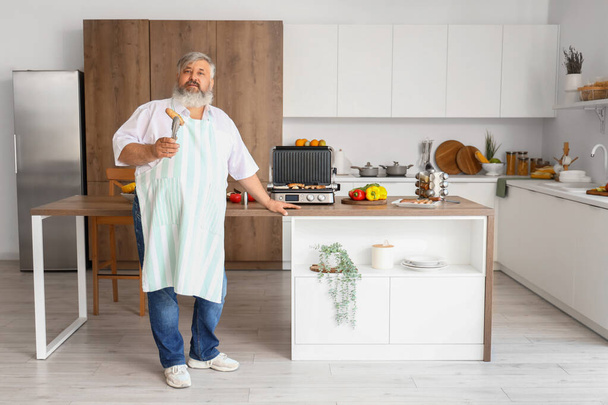 Homem maduro com tong e salsichas saborosas cozidas na moderna churrasqueira elétrica à mesa na cozinha - Foto, Imagem