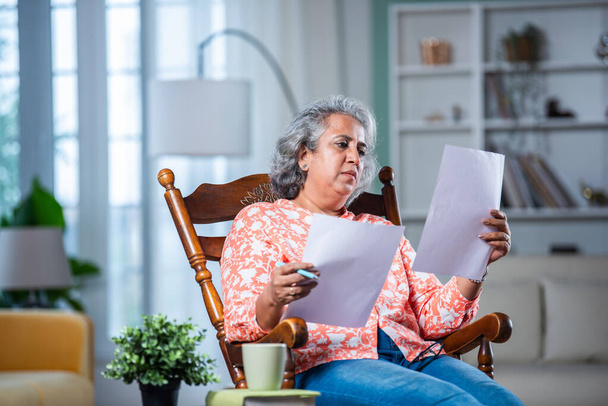 Domovská kancelář. Jistý indický asijské žena kontrola finanční zprávy zatímco sedí na houpací židli v obývacím pokoji - Fotografie, Obrázek