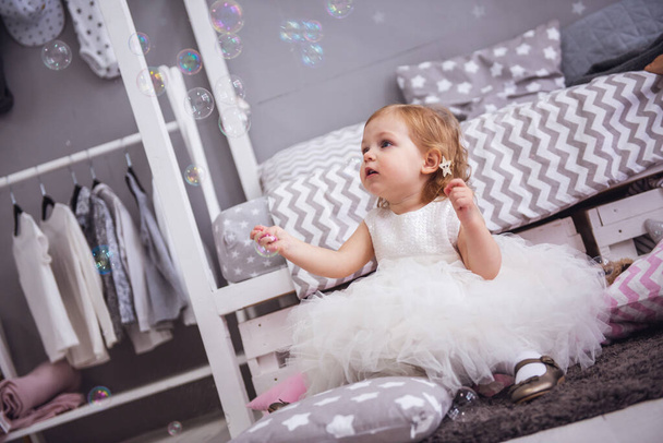 Cute little girl in fluffy white dress is playing with soap bubbles in the children's room - Photo, Image