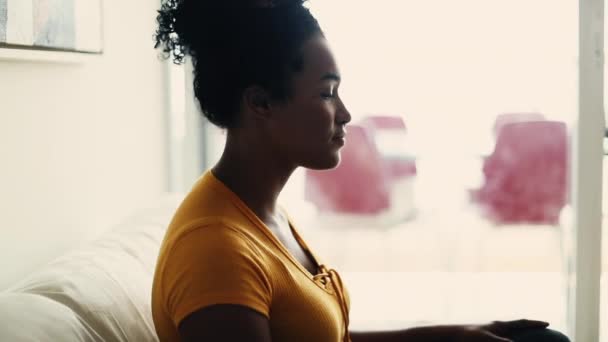 Perfil de una joven mujer negra sentada en el sofá de su casa en una tranquila soledad, contemplativa mirada de una persona afroamericana de 20 años - Imágenes, Vídeo