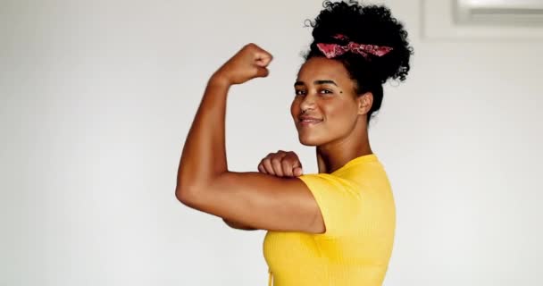 Счастливая уверенная молодая афроамериканка демонстрирует силу, сгибая руку, глядя в камеру. Одна черная латина показывает мышцы, мы можем сделать это символ, в желтой рубашке - Кадры, видео