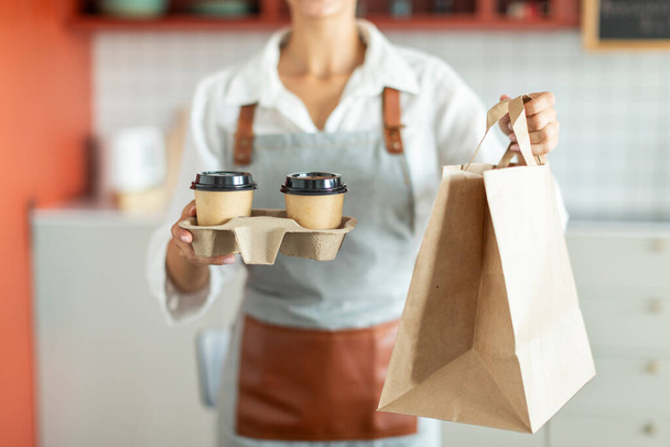 Closeup of waitress holding and serving paper cup of coffee and takeaway food in paper bag to customer in cafe, cropped shot - Photo, Image