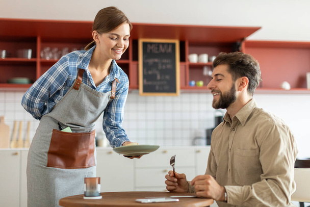 Alegre joven camarera que sirve un postre a feliz cliente masculino hambriento en la cafetería - Foto, imagen