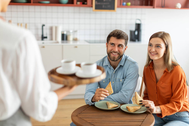 Kelnerka serwująca gorące aromatyczne napoje kawowe młodej parze, trzymająca tacę z kubkami, mężczyzna i kobieta goście czekający przy stoliku kawiarni - Zdjęcie, obraz