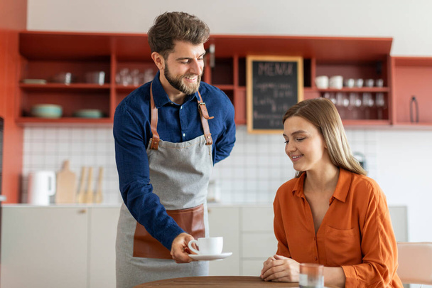 Start het concept van de eigenaar van een klein bedrijf. Vrolijke mannelijke ober serveert hete koffie aan lachende vrouwelijke klant, het geven van dame haar bestelling - Foto, afbeelding