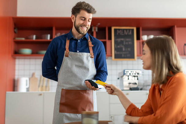 Happy male waiter holding payment terminal while lady client doing contactless purchase with her credit card in cafe - Photo, Image
