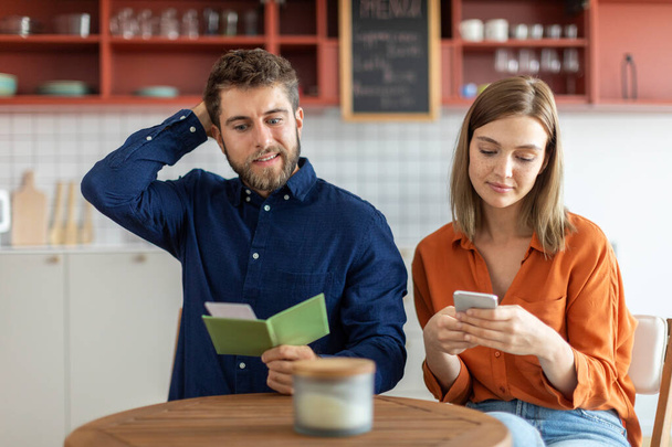 Schockierter Freund checkt Rechnung im Café und berührt Kopf, bezahlt Mittagessen, während seine Freundin Handy benutzt - Foto, Bild