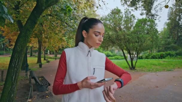 Спортивна жінка перевіряє розумний годинник на зелені парку, готуючись до фітнес-рутингу крупним планом. Фокусований бігун налаштування цифрових пристроїв перед тренуванням на осінній природі. Щоденний моніторинг здоров'я за технологіями - Кадри, відео