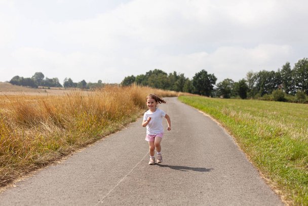 Little girl happily running along a quiet rural road, surrounded by the beauty and tranquility of the countryside - Photo, Image