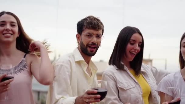 Skupina nadšených mladých tisíciletých přátel držících červené sklenice víno tančící společně na letním venkovním prostranství. Veselá skupina šťastní lidé těší západ slunce alkoholické nápoje na střešní party  - Záběry, video