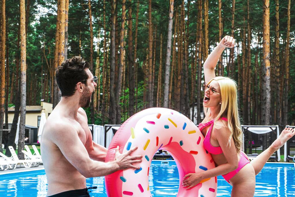 Smiling man and woman enjoying weekend outdoor and having fun while holding inflatable ring near swimming pool. Summer vacation, active leisure, lifestyle concept - Photo, Image