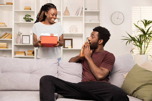 Surprised Man Receiving Gift From Woman At Home. Joyful African American Couple Sharing A Happy Moment With A Present. Excitement, Celebration, And Relationship Concept. - Zdjęcie, obraz