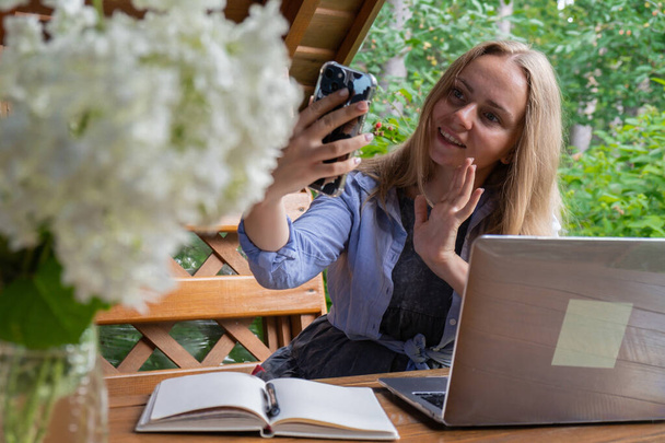 Nuori onnellinen nainen tekee videopuhelun älypuhelimella puisessa alkovissa. Rento ulkoilu korostaa mukavuutta ja tuottavuutta. Etätyön oppimisen käsite - Valokuva, kuva