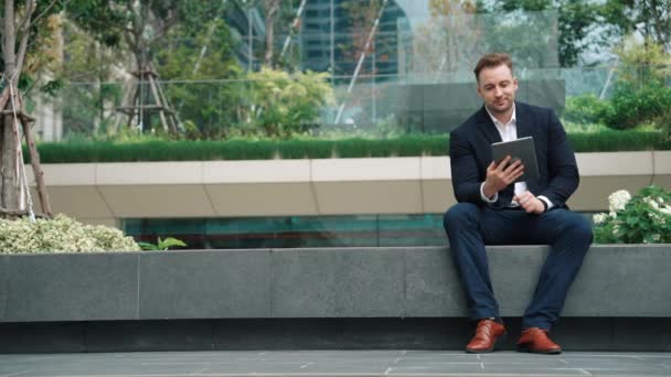 Профессиональный бизнесмен сидит в парке во время работы над планшетом, читая отчет от маркетинговой команды. Счастливый менеджер смотрит на увеличение продаж или успешный проект, улыбаясь при этом экогороду. Урбан - Кадры, видео