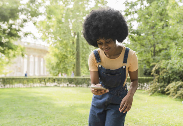 Eine lebhafte junge Frau, die inmitten der ruhigen Kulisse eines sonnenbeschienenen Parks mit ihrem Handy beschäftigt ist. - Foto, Bild
