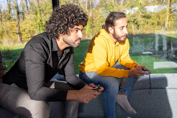 Two friends are deeply focused while playing video games in a living room filled with natural light. The intensity on their faces reflects the immersion in the game, with game controllers in hand. The - Photo, Image