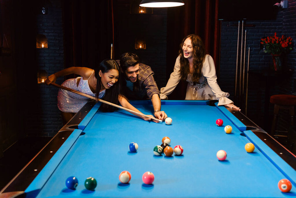 Young diversity group of people playing pool together with smile, enjoyment and fun. Young people spend time in billiards room at the nightclub. Men, women friends playing billiards. Nightlife concept - Photo, Image