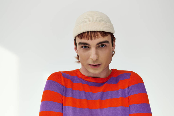 позитивный модный гей в белой шляпе в ярком одеянии позирует на сером фоне и смотрит в камеру - Фото, изображение