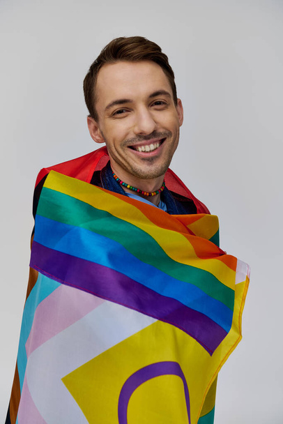 atraente alegre gay homem no vibrante casual traje segurando arco-íris bandeira e sorrindo para câmera - Foto, Imagem