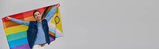 allegro attraente gay uomo in vibrante casual abbigliamento tenendo arcobaleno bandiera e sorridente a fotocamera banner - Foto, immagini