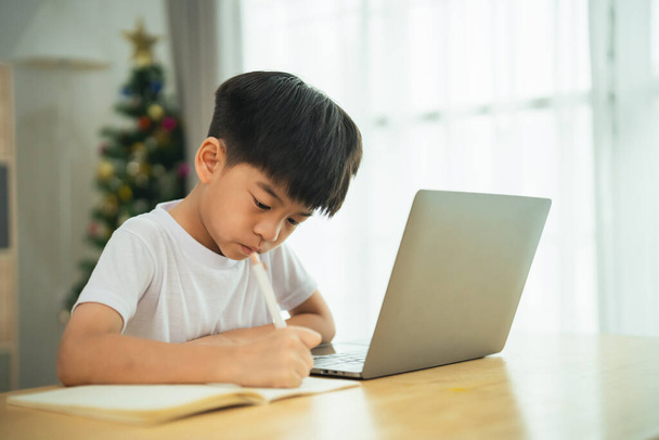 Een jonge jongen zit aan een tafel met een laptop en een potlood. Hij schrijft iets op een stuk papier. - Foto, afbeelding