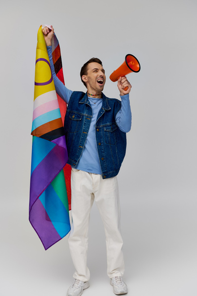 νέος όμορφος ομοφυλόφιλος άνδρας με ζεστή ενδυμασία κρατώντας τη σημαία του ουράνιου τόξου και χρησιμοποιώντας μεγάφωνο σε γκρι φόντο - Φωτογραφία, εικόνα