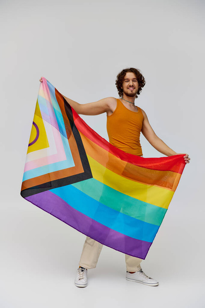 カメラで虹の旗を保持し,喜んで微笑んでいる暗い髪の肯定的な魅力的なゲイ男性 - 写真・画像