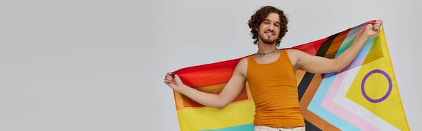 hilpeästi houkutteleva homo mies tummilla hiuksilla pitäen sateenkaaren lippua ja hymyillen onnellisesti kameralle, banneri - Valokuva, kuva
