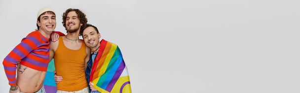 three jolly gay men in cozy clothing posing actively with rainbow flag on gray backdrop, banner - Фото, изображение