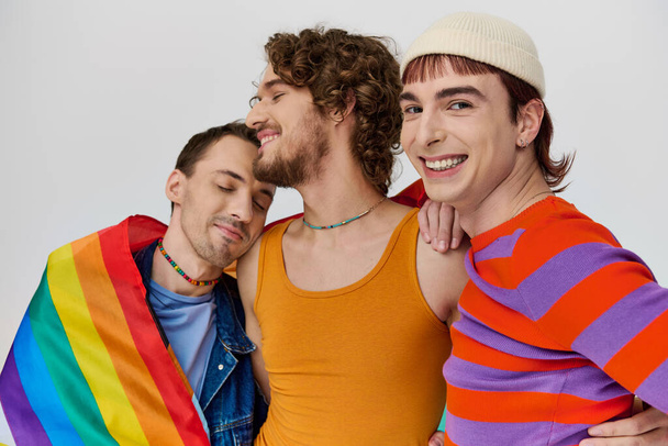 three joyful stylish gay men in cozy clothing posing actively with rainbow flag on gray backdrop - Photo, Image
