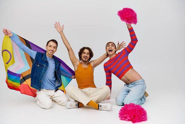 gioioso attraente gay gli uomini in vibrante vestiti posa con arcobaleno bandiera e pon pon su grigio sfondo - Foto, immagini