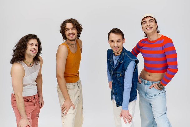 τέσσερις χαρούμενους και εμφανίσιμους γκέι άντρες με έντονα ρούχα να χαμογελούν στην κάμερα ενώ ποζάρουν σε γκρι φόντο - Φωτογραφία, εικόνα