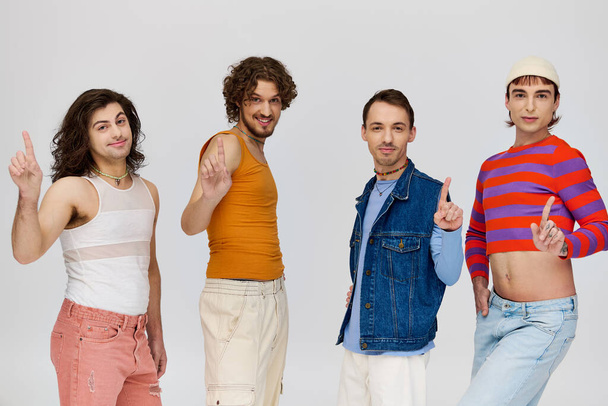 vier positieve goed uitziende gay mannen in levendige kleding glimlachen op camera terwijl poseren op grijze achtergrond - Foto, afbeelding