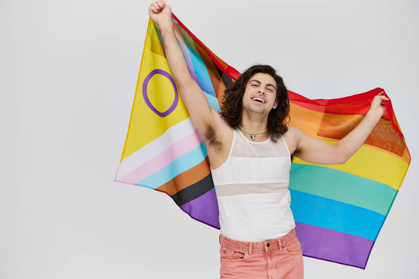 aantrekkelijk vrolijke gay man met lang donker haar poseren met regenboog vlag en kijken naar camera - Foto, afbeelding