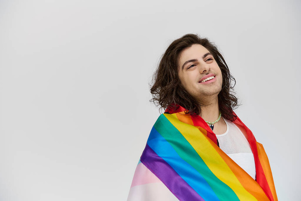 красивый модный веселый гей в яркой одежде с радужным флагом, смотрящий на серый фон - Фото, изображение