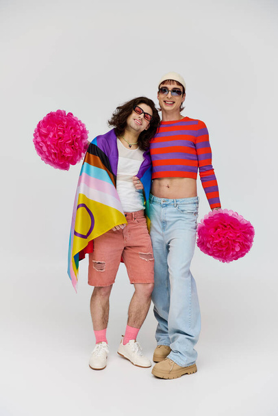 dva radostné svůdné gayové v tučných oděvech se slunečními brýlemi pózující s duhovou vlajkou a pom poms - Fotografie, Obrázek