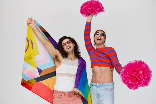 two joyful alluring gay men in bold attires with sunglasses posing with rainbow flag and pom poms - Photo, Image