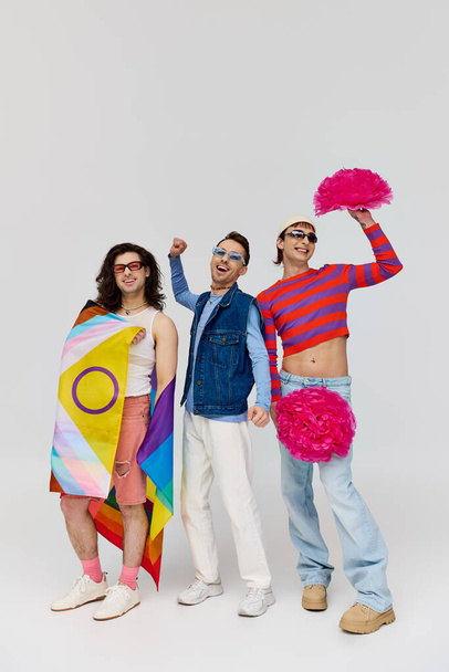 drie vrolijke stijlvolle gay mannen in vet kleding met zonnebril poseren met pom poms en regenboog vlag - Foto, afbeelding